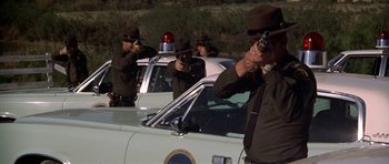 Movie still from “The Sugarland Express” (1974), directed by Steven Spielberg – A group of police officers standing next to each other on the side of the road; Wide shot, Over the shoulder angle