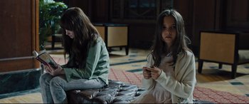 Movie still from “The Super” (2017), directed by Stephan Rick – Two young girls sitting on a bench in a living room; Medium shot, High angle