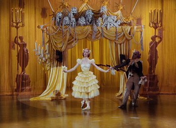 Movie still from “The Tales of Hoffmann” (1951), directed by Michael Powell – A woman in a yellow dress is standing in front of a chandelier; Wide shot, Low angle