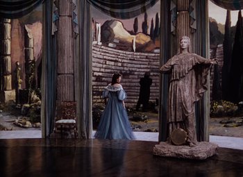 Movie still from “The Tales of Hoffmann” (1951), directed by Michael Powell – A woman in a blue dress standing next to a statue; Wide shot, Over the shoulder angle