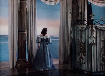 Movie still from “The Tales of Hoffmann” (1951), directed by Michael Powell – A woman in a blue dress standing in front of an open door; Wide shot, High angle
