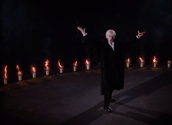 Movie still from “The Tales of Hoffmann” (1951), directed by Michael Powell – An old man in a black coat standing in front of a row of lit candles; Wide shot, Low angle
