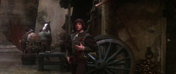Movie still from “The Taming of The Shrew” (1967), directed by Franco Zeffirelli – A man dressed in medieval clothing standing next to a wagon wheel; Medium shot, Over the shoulder angle