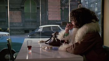 Movie still from “The Tenant” (1976), directed by Roman Polanski – A person sitting at a table with a drink; Medium shot, Over the shoulder angle