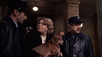 Movie still from “The Tenant” (1976), directed by Roman Polanski – A woman holding a brown dog in front of a group of men; Medium shot, Low angle