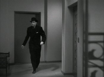 Movie still from “The Thin Man” (1934), directed by W.S. Van Dyke – A man in a black suit and a hat walking in a hallway; Wide shot, High angle