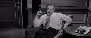 Movie still from “The Three Faces of Eve” (1957), directed by Nunnally Johnson – A man sitting in a chair smoking a cigarette; Medium shot, High angle