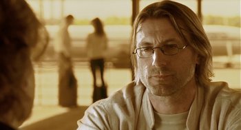 Movie still from “The Ugly Swans” (2006), directed by Konstantin Lopushanskiy – A person wearing glasses with people in the background; Close Up shot, Over the shoulder angle