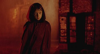 Movie still from “The Ugly Swans” (2006), directed by Konstantin Lopushanskiy – A child in a red hooded cloak stands in a dark room; Medium shot, Over the shoulder angle