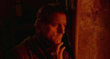 Movie still from “The Ugly Swans” (2006), directed by Konstantin Lopushanskiy – A man with long black hair is leaning against a wall with his hand under his chin; Close Up shot, Low angle