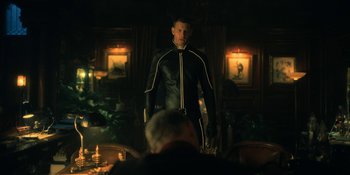 Movie still from “The Umbrella Academy” (2019), created by Steve Blackman – A man in a black and yellow suit standing in front of a table; Medium shot, Over the shoulder angle