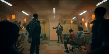 Movie still from “The Umbrella Academy” (2019), created by Steve Blackman – A group of people sitting in a room with a man in a business suit; Wide shot, Over the shoulder angle