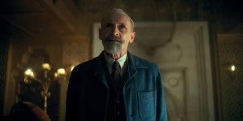Movie still from “The Umbrella Academy” (2019), created by Steve Blackman – An old man with a beard wearing a suit and tie; Medium shot, Low angle