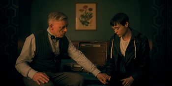 Movie still from “The Umbrella Academy” (2019), created by Steve Blackman – A man sitting next to a young boy in front of a suitcase; Medium shot, Over the shoulder angle