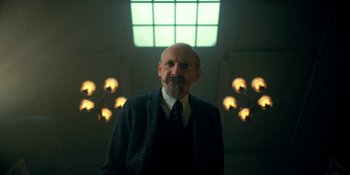 Movie still from “The Umbrella Academy” (2019), created by Steve Blackman – An older man in a suit and tie in front of lights; Medium shot, Low angle