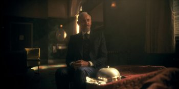 Movie still from “The Umbrella Academy” (2019), created by Steve Blackman – An older man in a suit sitting in front of a silver platter; Medium shot, Over the shoulder angle