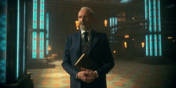 Movie still from “The Umbrella Academy” (2019), created by Steve Blackman – An older man in a suit and tie holding a book; Medium shot, Low angle