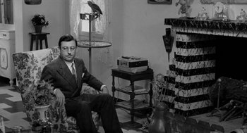 Movie still from “The Visit” (1964), directed by Bernhard Wicki – A man in a suit sitting on a chair in a room; Medium shot, Low angle