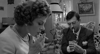 Movie still from “The Visit” (1964), directed by Bernhard Wicki – A man and a woman are sitting in a living room smoking; Medium shot, Low angle