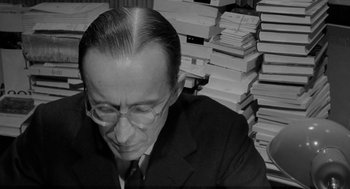 Movie still from “The Visit” (1964), directed by Bernhard Wicki – A man in a suit and glasses sitting in front of a stack of books; Medium shot, High angle