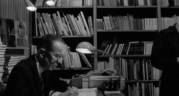 Movie still from “The Visit” (1964), directed by Bernhard Wicki – An older man sitting at a desk in front of a book shelf; Medium shot, High angle