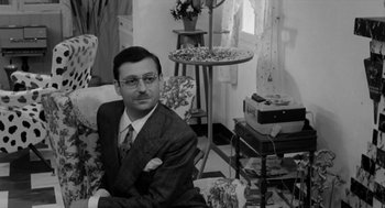Movie still from “The Visit” (1964), directed by Bernhard Wicki – A black and white photo of a man sitting on a couch; Medium shot, Over the shoulder angle
