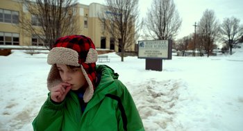 Movie still from “The Visit” (2015), directed by M. Night Shyamalan – A boy in a green jacket and a red and black hat is standing in the snow; Medium shot, High angle