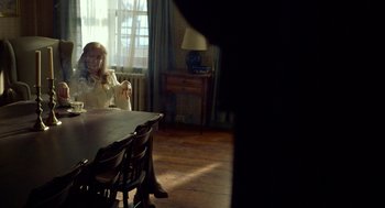 Movie still from “The Visit” (2015), directed by M. Night Shyamalan – A woman sitting at a dining room table in front of a lamp; Medium shot, Over the shoulder angle
