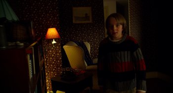 Movie still from “The Visit” (2015), directed by M. Night Shyamalan – A young boy standing in a living room with a lamp; Medium shot, Over the shoulder angle