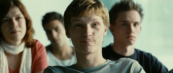 Movie still from “The Wave” (2008), directed by Dennis Gansel – A young man is smiling for the camera while two other young men are standing behind him; Close Up shot, Low angle