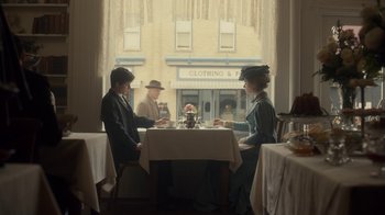 Movie still from “Anne with an E” (2017), created by Moira Walley-Beckett – Two people sitting at a table in front of a store window; Wide shot, Over the shoulder angle