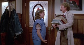 Movie still from “The Woman in Red” (1984), directed by Gene Wilder – A young girl standing in front of an older man holding a stack of books; Medium shot, Over the shoulder angle