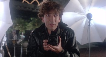 Movie still from “The Woman in Red” (1984), directed by Gene Wilder – A man in a black jacket holding up a camera; Close Up shot, Low angle