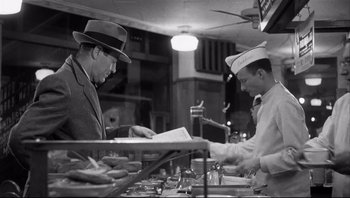 Movie still from “The Wrong Man” (1956), directed by Alfred Hitchcock – A man in a suit and hat is looking at a menu in front of another man in a chef's hat; Medium shot, Over the shoulder angle
