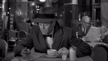 Movie still from “The Wrong Man” (1956), directed by Alfred Hitchcock – A black and white photo of a man sitting at a table drinking a cup of coffee; Medium shot, Over the shoulder angle