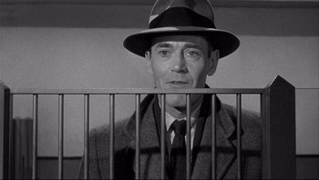 Movie still from “The Wrong Man” (1956), directed by Alfred Hitchcock – A man wearing a suit and a hat behind a fence; Medium shot, Low angle