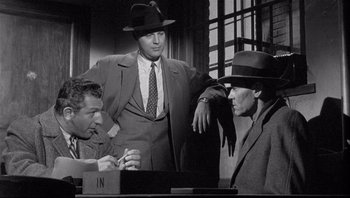 Movie still from “The Wrong Man” (1956), directed by Alfred Hitchcock – A man in a suit and a hat standing next to two other men; Medium shot, Over the shoulder angle