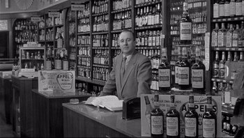 Movie still from “The Wrong Man” (1956), directed by Alfred Hitchcock – A man standing behind a counter in a liquor store; Medium shot, Low angle