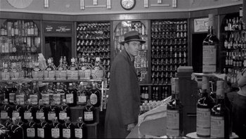 Movie still from “The Wrong Man” (1956), directed by Alfred Hitchcock – A black and white photo of a man standing in a liquor store; Wide shot, High angle