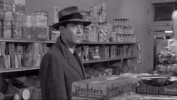 Movie still from “The Wrong Man” (1956), directed by Alfred Hitchcock – A man standing in front of a shelf filled with cans; Medium shot, Over the shoulder angle