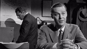 Movie still from “The Wrong Man” (1956), directed by Alfred Hitchcock – A man sitting at a desk in front of another man in a suit; Medium shot, Over the shoulder angle