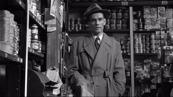 Movie still from “The Wrong Man” (1956), directed by Alfred Hitchcock – A man in a trench coat and hat standing in front of shelves; Medium shot, Over the shoulder angle