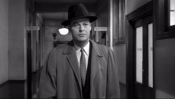 Movie still from “The Wrong Man” (1956), directed by Alfred Hitchcock – A black and white photo of a man wearing a hat and a suit; Medium shot, Over the shoulder angle