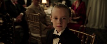 Movie still from “The Young and Prodigious T.S. Spivet” (2013), directed by Jean-Pierre Jeunet – A young boy in a tuxedo sitting in a chair in front of other people; Close Up shot, Over the shoulder angle