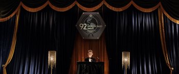 Movie still from “The Young and Prodigious T.S. Spivet” (2013), directed by Jean-Pierre Jeunet – A man standing at a podium in front of a sign; Wide shot, High angle