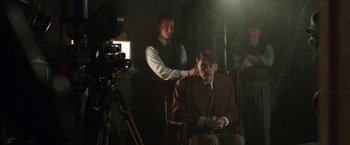 Movie still from “Their Finest” (2016), directed by Lone Scherfig – A man sitting in a chair in front of a movie camera; Medium shot, Over the shoulder angle