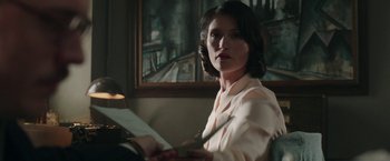 Movie still from “Their Finest” (2016), directed by Lone Scherfig – A woman sitting in front of a painting holding papers; Medium shot, Over the shoulder angle