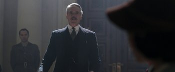 Movie still from “Their Finest” (2016), directed by Lone Scherfig – An older man in a suit and tie in a dark room; Medium shot, Over the shoulder angle