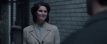 Movie still from “Their Finest” (2016), directed by Lone Scherfig – A woman wearing a coat and hat standing in front of a brick wall; Close Up shot, Over the shoulder angle