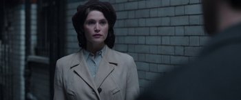 Movie still from “Their Finest” (2016), directed by Lone Scherfig – A woman wearing a coat and hat standing next to a brick wall; Close Up shot, Over the shoulder angle